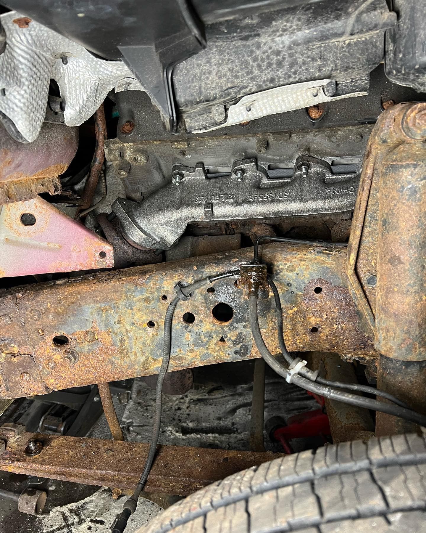 Rusty vehicle frame with undercarriage components, including hoses and a tire visible.