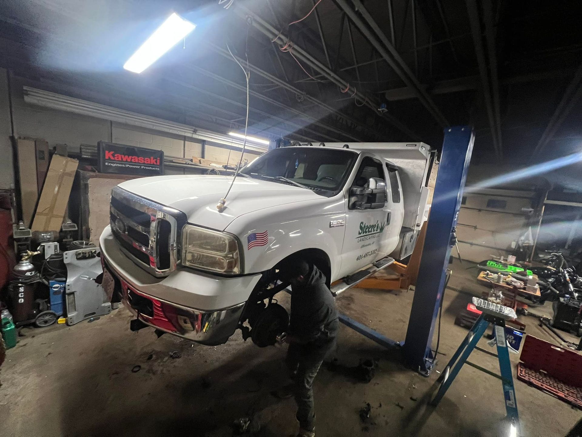 A white truck raised on a lift in a repair shop; a person works beneath it, likely repairing brakes.