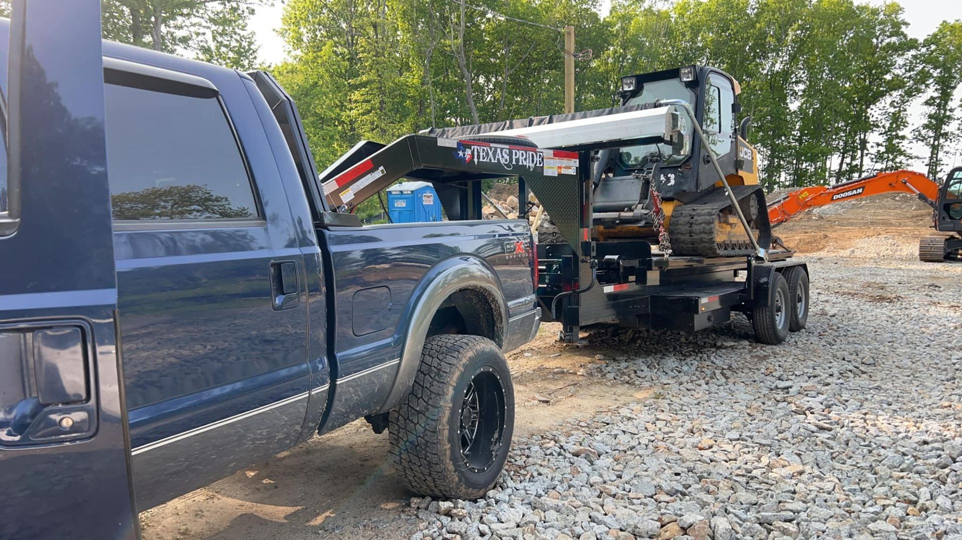 Blue truck towing a trailer with a piece of heavy construction equipment on gravel.