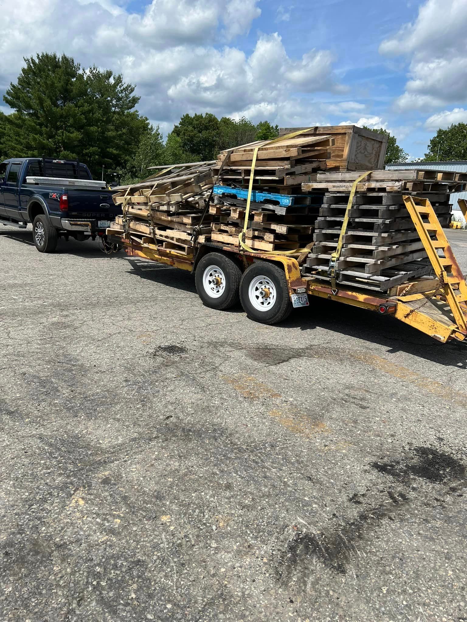 Black pickup truck towing a trailer loaded with wooden pallets on a gravel lot under a cloudy sky.