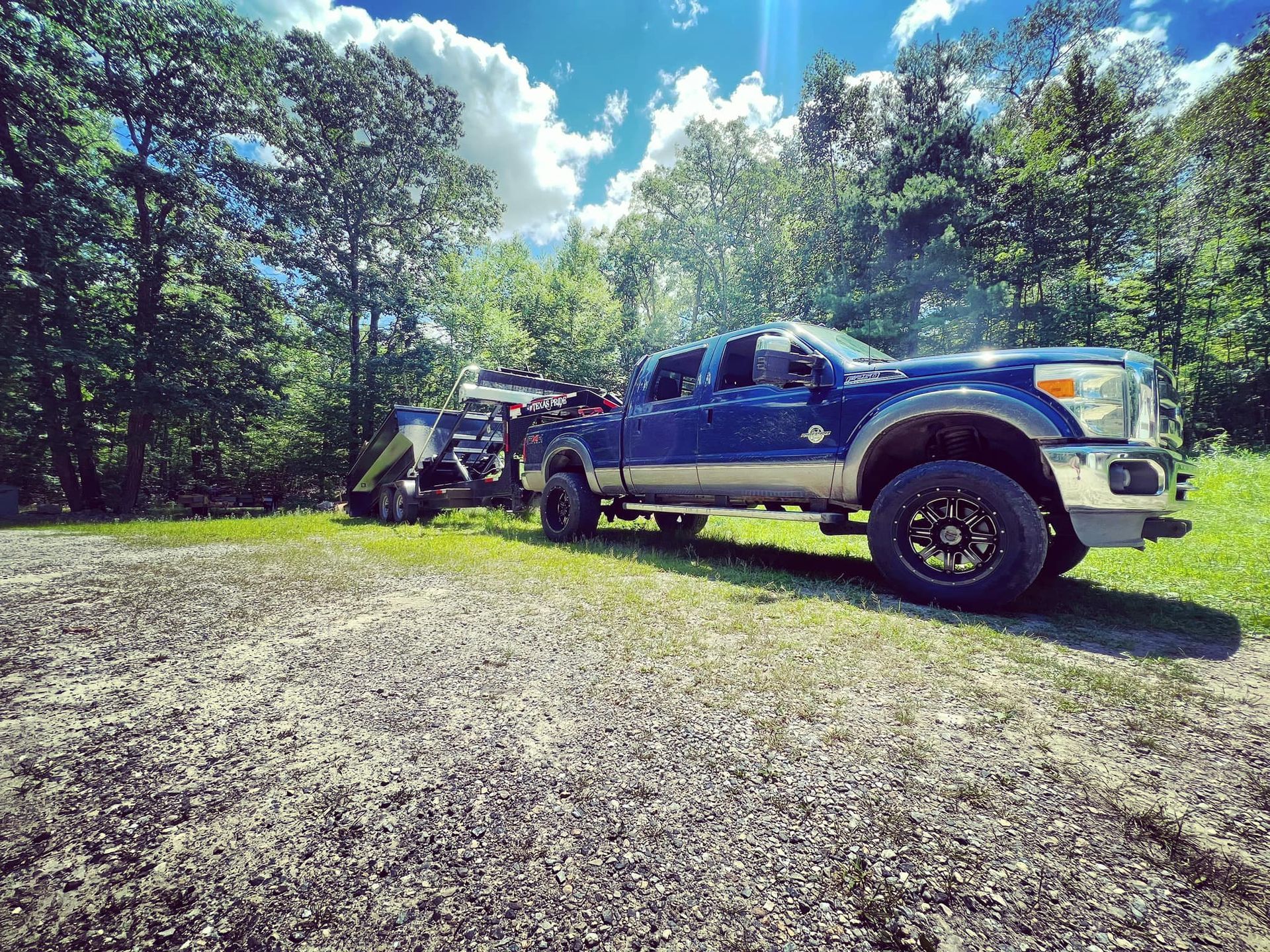Blue truck towing trailer on gravel road; trees and sky in background.