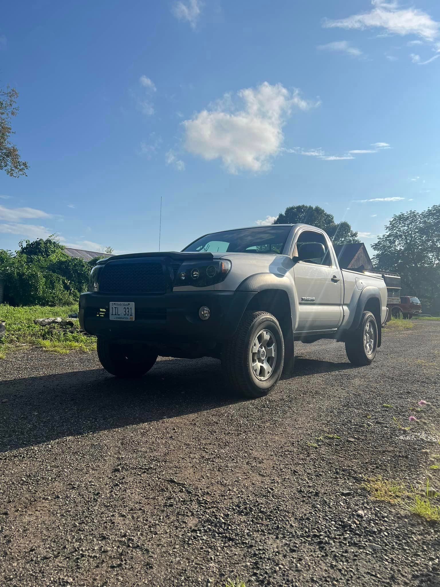 Gray Toyota Tacoma pickup truck parked on gravel under a blue sky with clouds.