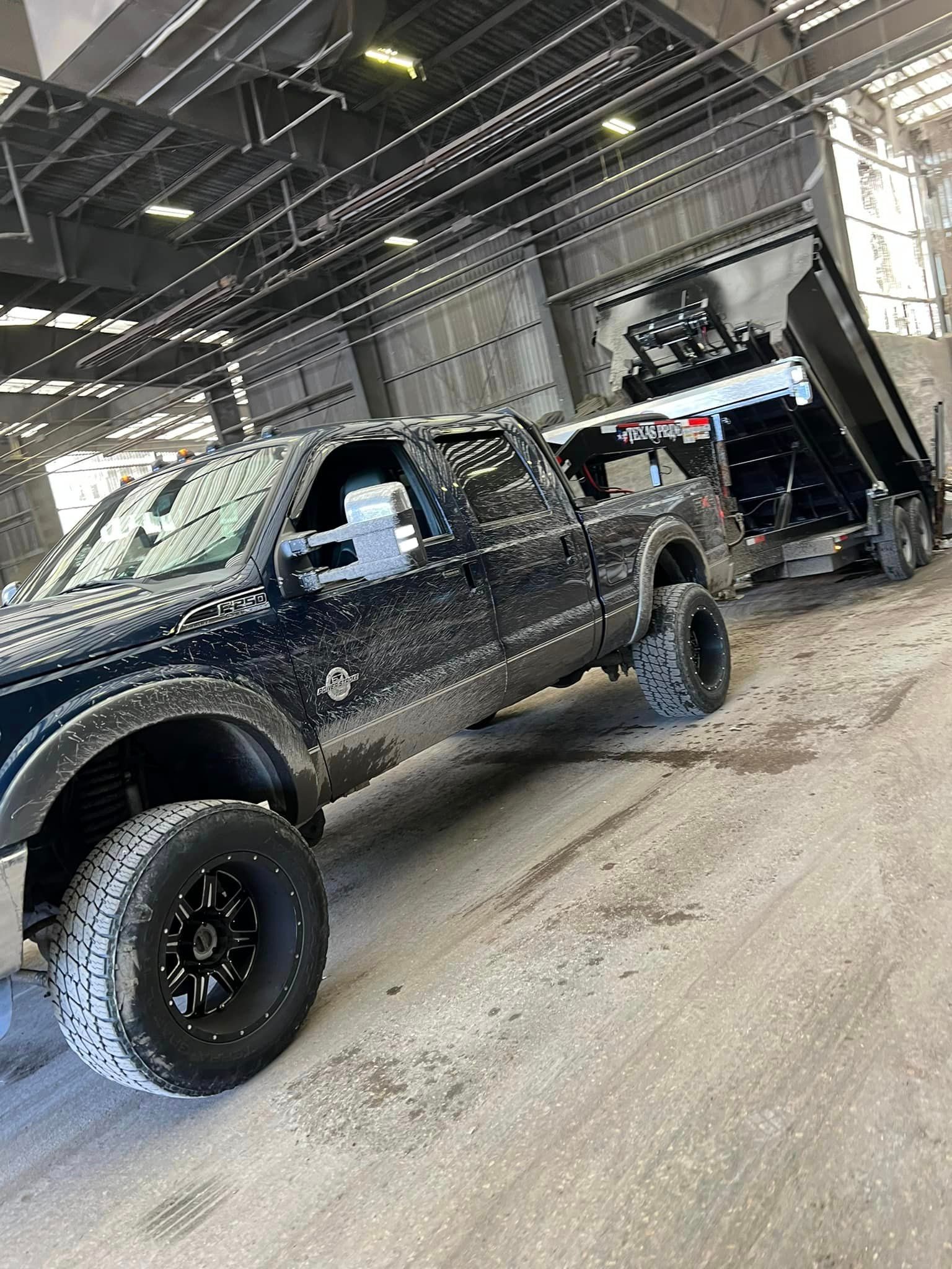 Dark pickup truck with lifted wheels pulling a dump trailer, both covered in grime, in a commercial building.