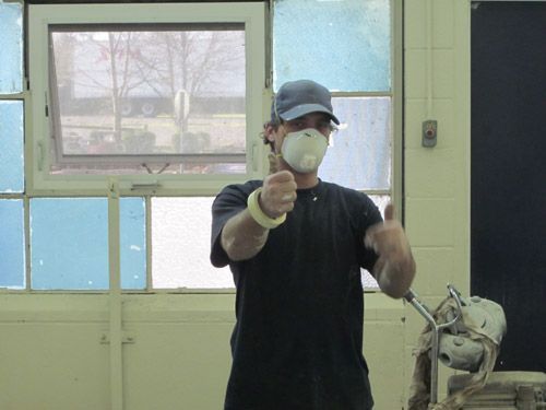 A man wearing a mask is giving a thumbs up in front of a window.