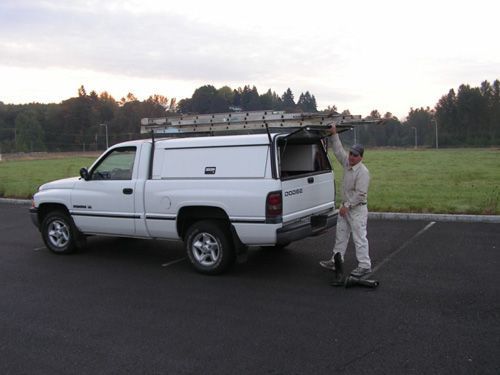 A man is standing next to a white truck with a ladder on top of it.