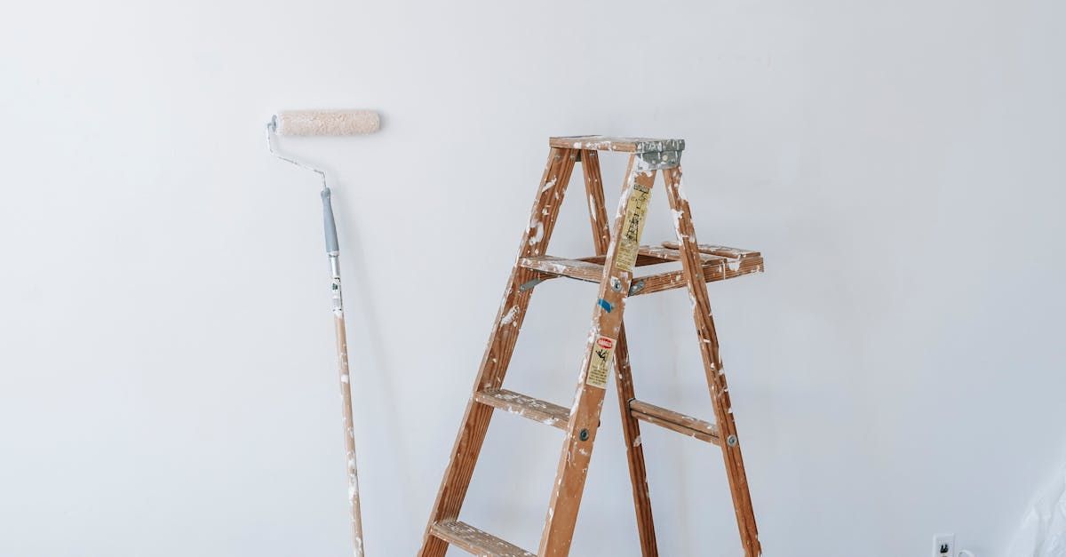 A wooden ladder is sitting next to a paint roller on a white wall.