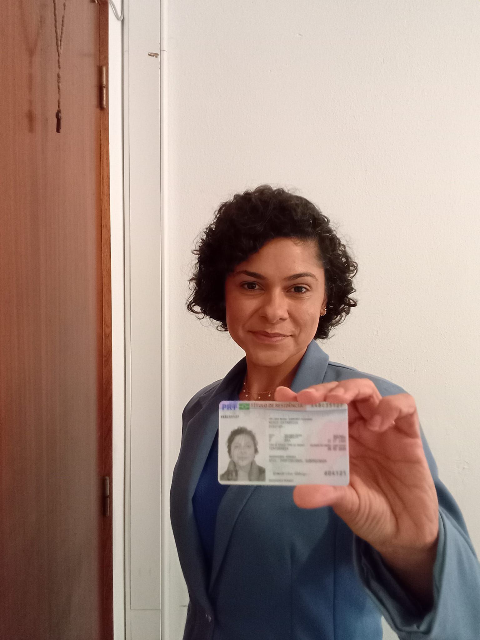 A person in a blue blazer holds up a government-issued identification card in front of a white wall and a brown door.