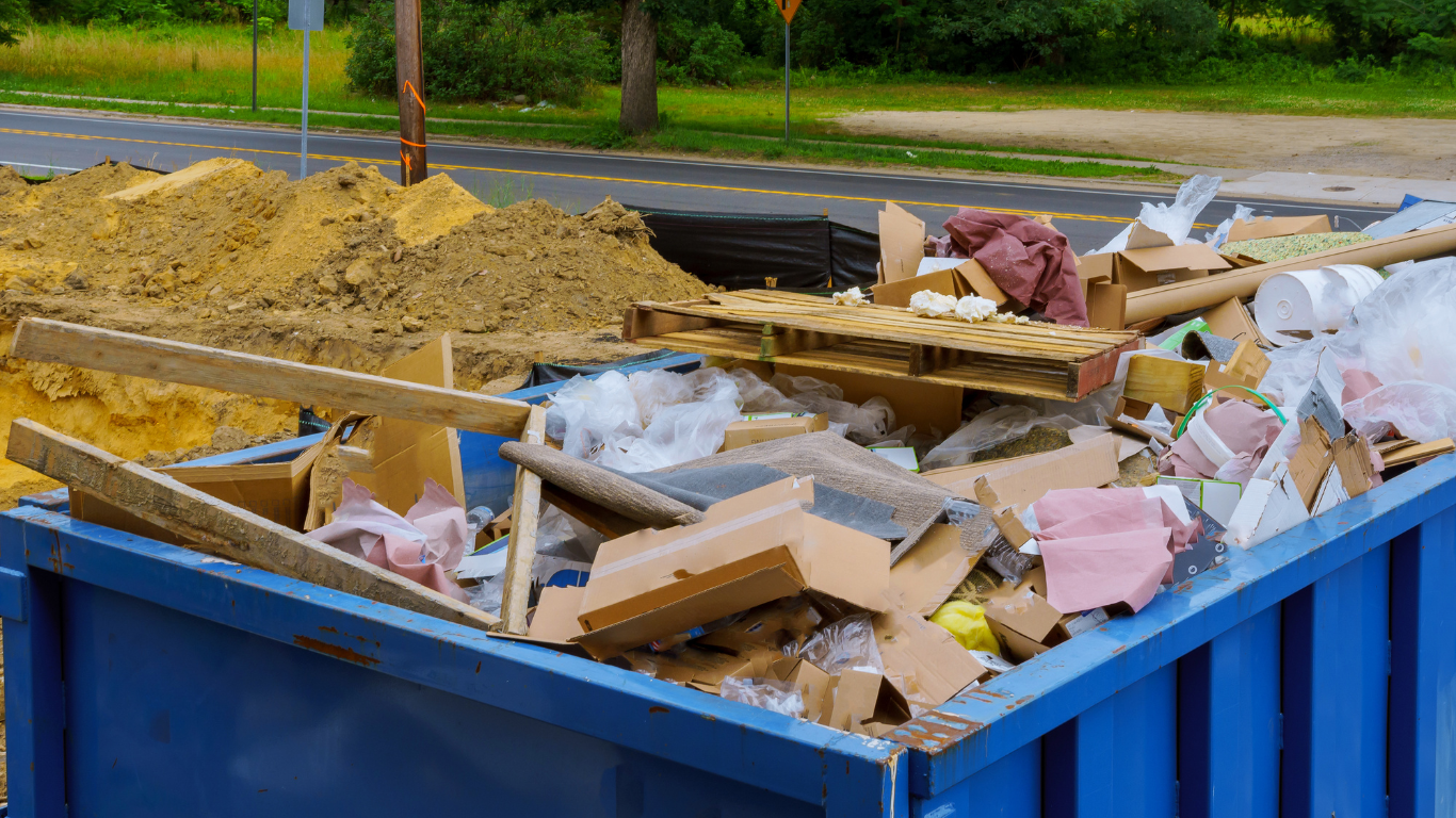 A blue dumpster filled with lots of trash is sitting next to a pile of dirt.