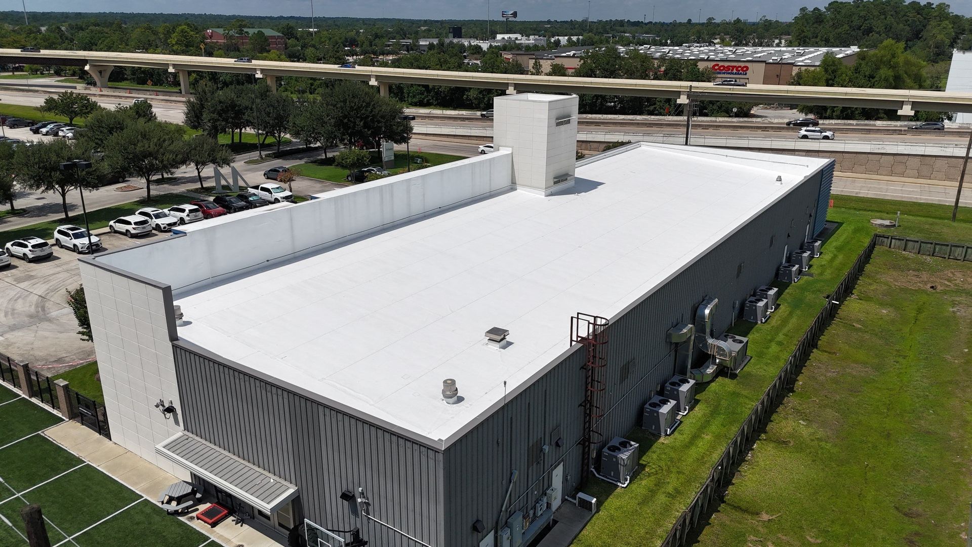 Large, light-colored industrial building with a flat roof. A tower rises at one end. Cars parked nearby. Green grass in the foreground.