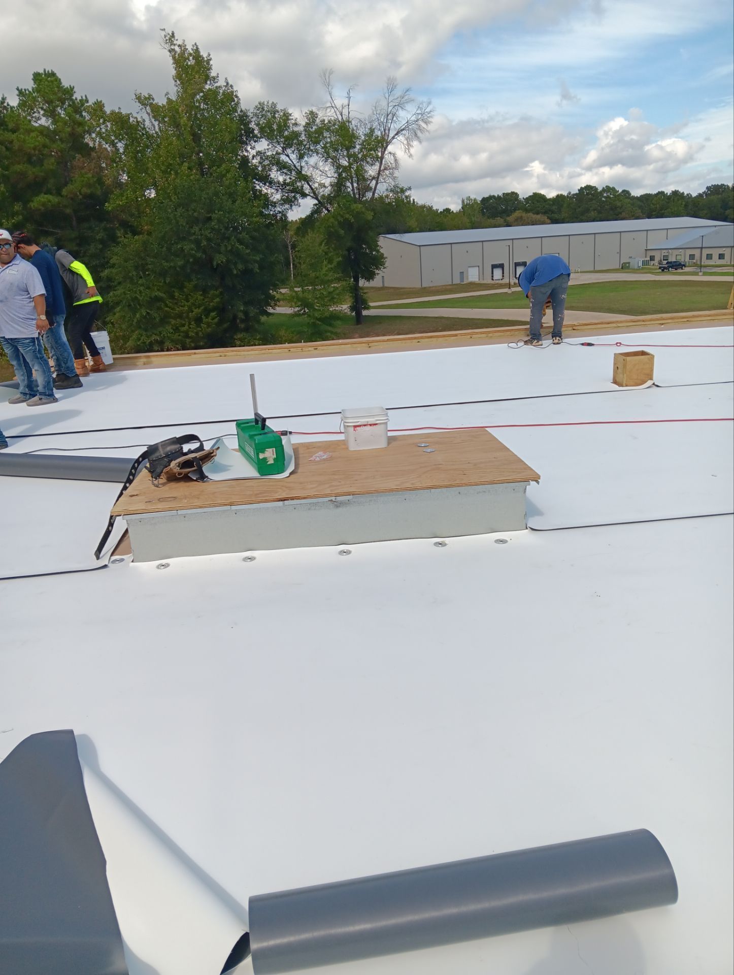 Construction workers on a white roof, installing material with tools and a heat gun. Cloudy sky.