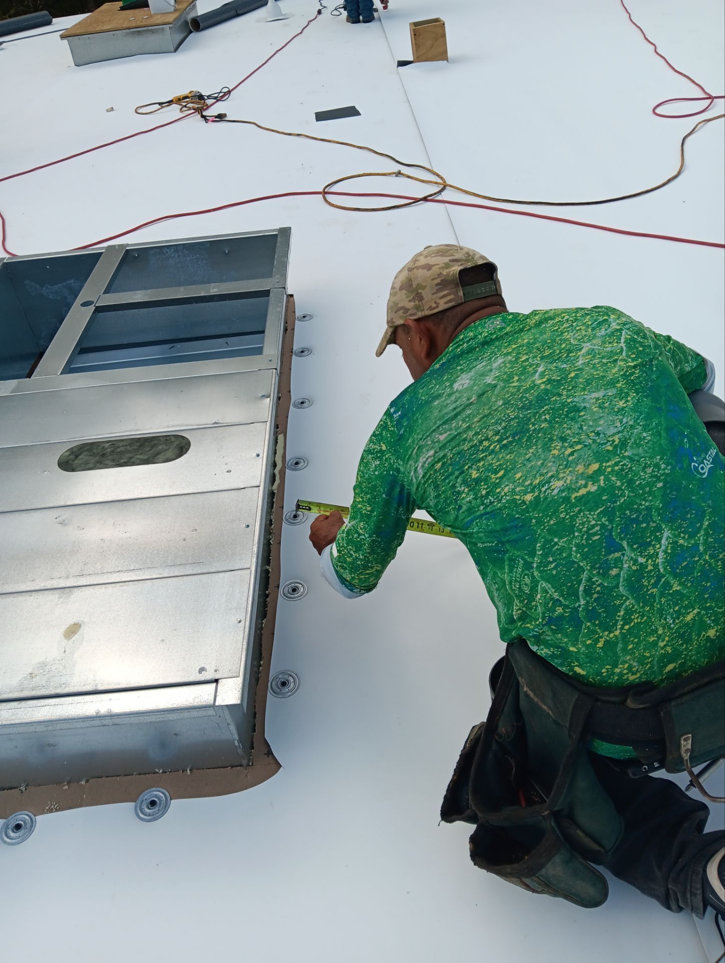 Roofer in green shirt measuring a metal structure on a white flat roof. Red cord and tools visible.