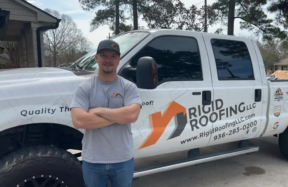 Man in grey shirt, arms crossed, stands next to a white Rigid Roofing truck, logo visible.