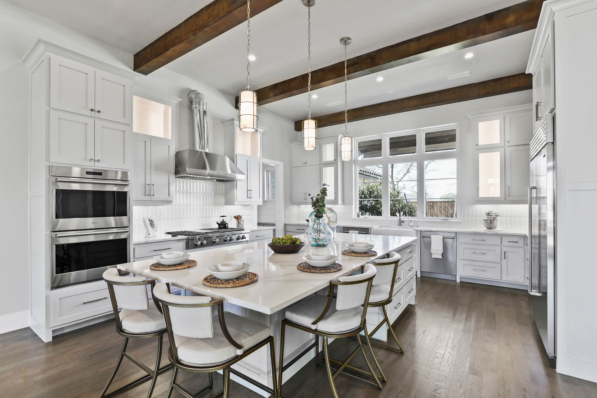  A modern kitchen in a custom-built home with white cabinets, stainless steel appliances, a central island with seating, pendant lights, and wooden beams on the ceiling.
