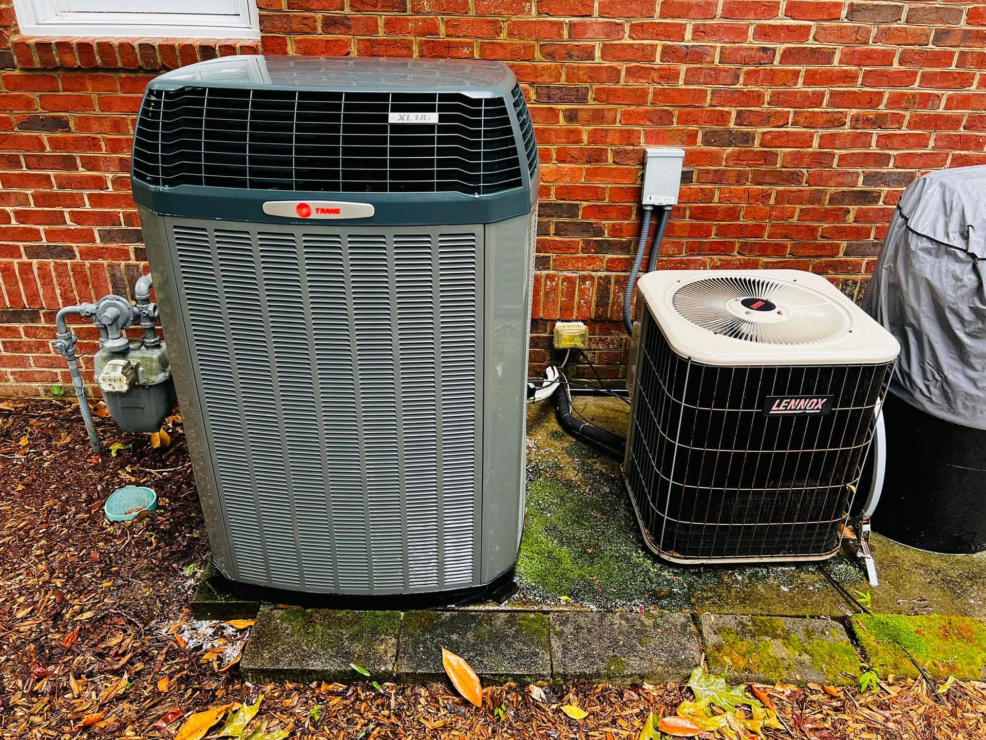 Two air conditioning units, Trane and York, next to a brick wall.