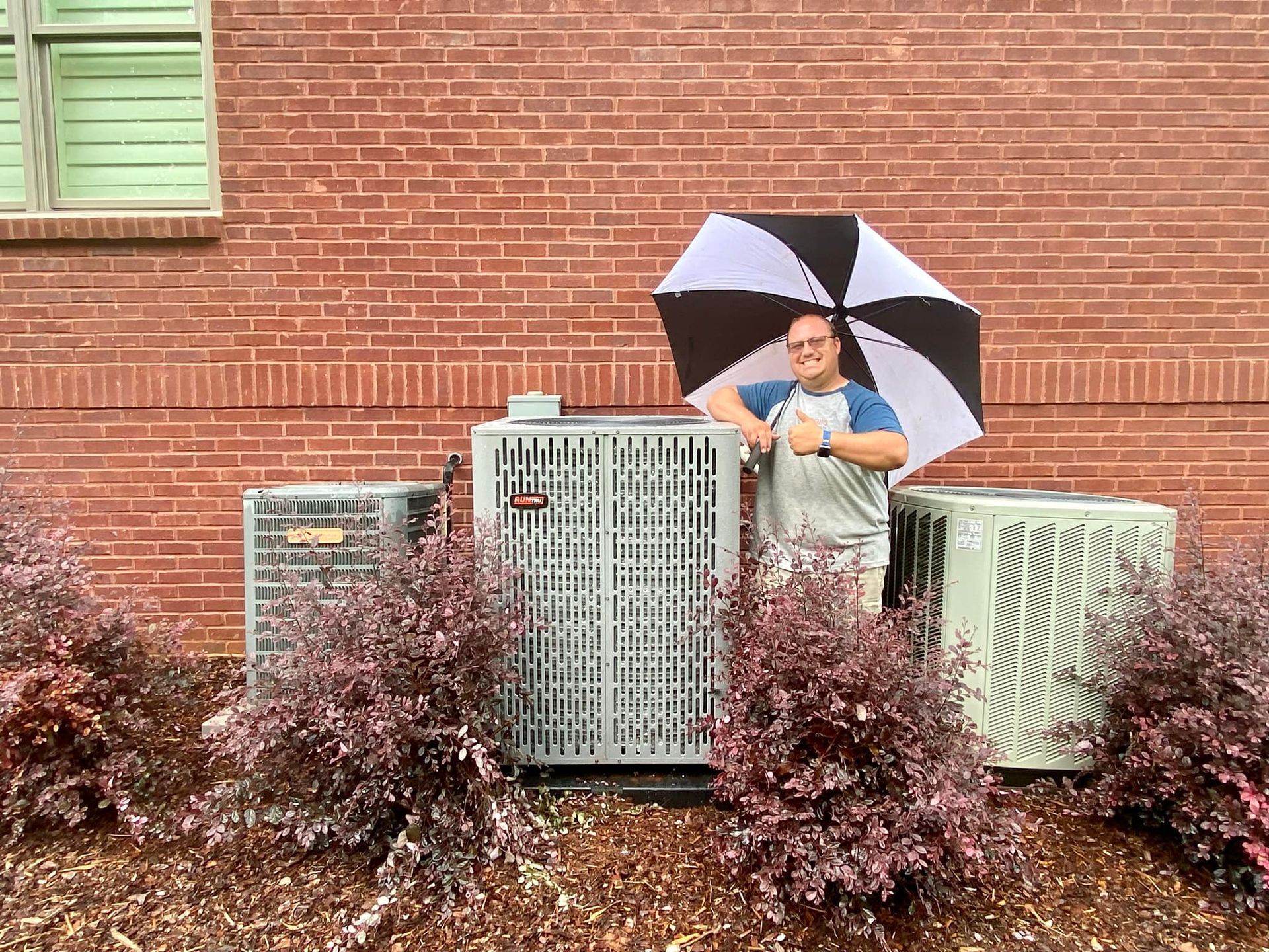 Person with umbrella stands by air conditioning units next to a brick building.
