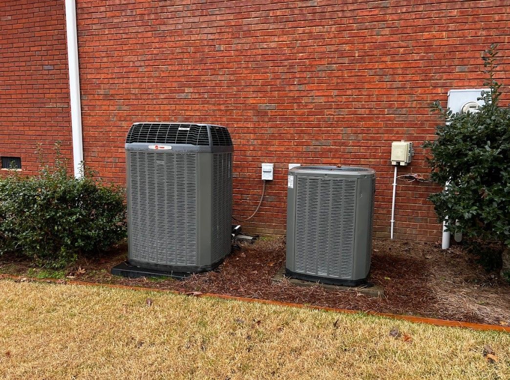 Two outdoor HVAC units against a brick wall, grass in the foreground.