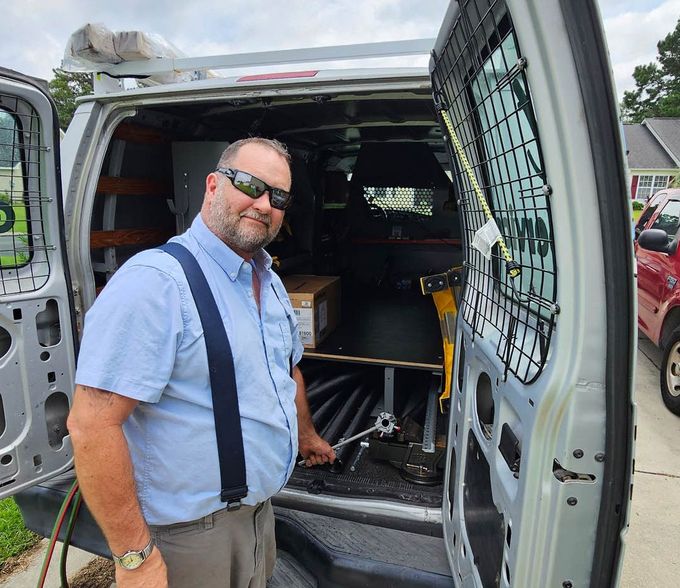 Man in sunglasses stands in back of a van holding a tool, smiling. The van's side doors are open, showing tools and equipment inside.