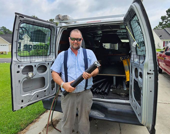 Man in work clothes stands by an open service van holding a black pipe. He is in front of a residential setting.