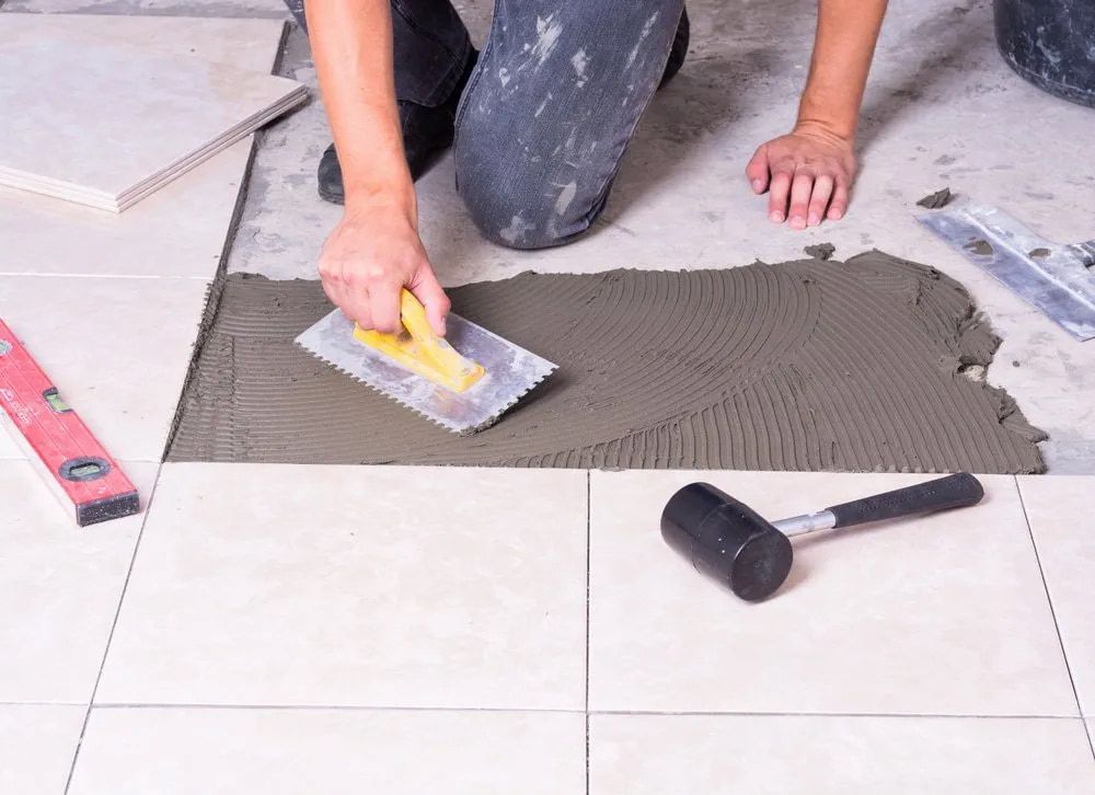 A Person Is Laying Tiles On The Floor With A Trowel — Corrimal Ceramic Tiles Pty Ltd in Gerringong, NSW