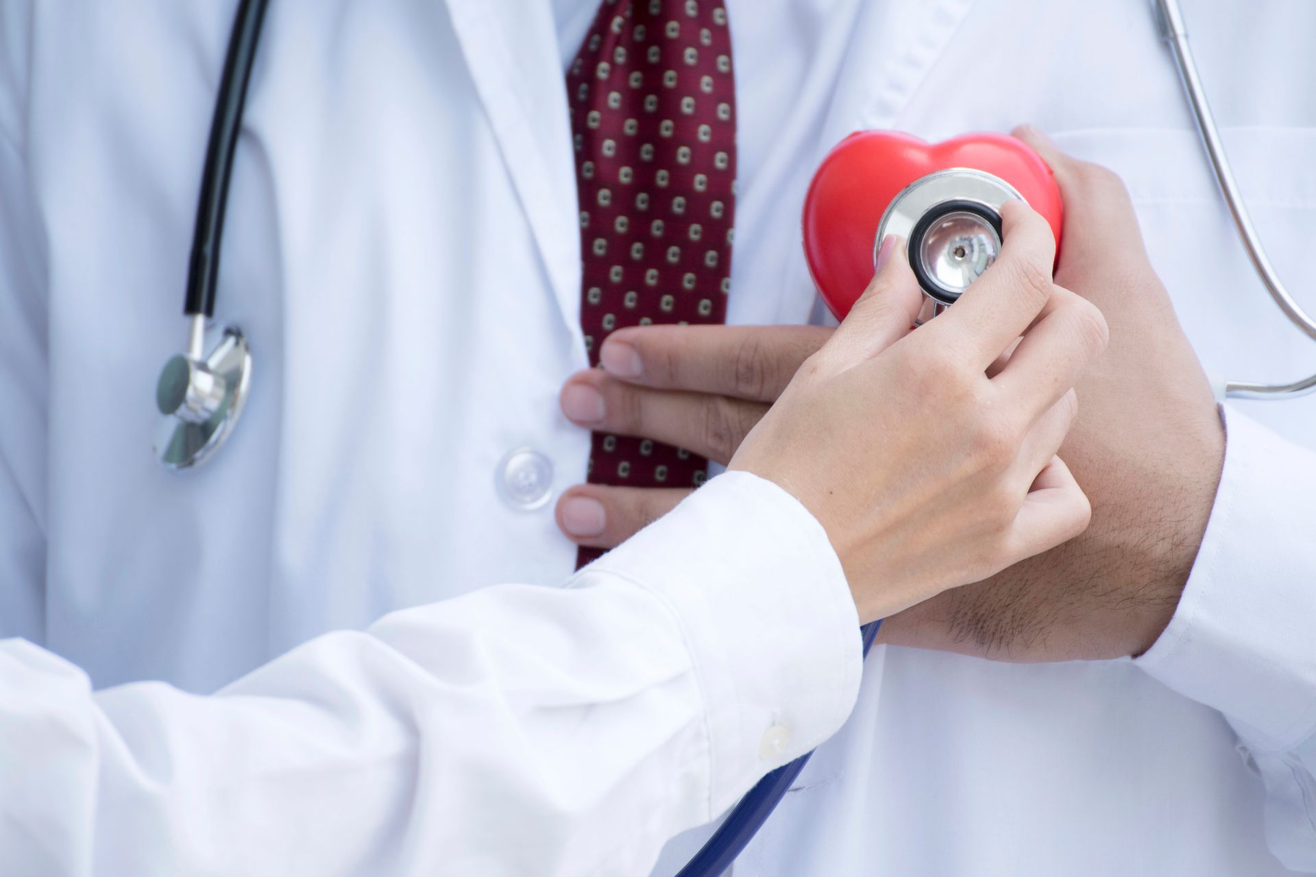 Un operatore sanitario utilizza uno stetoscopio per auscultare un piccolo modello di cuore rosso appoggiato su un camice bianco.