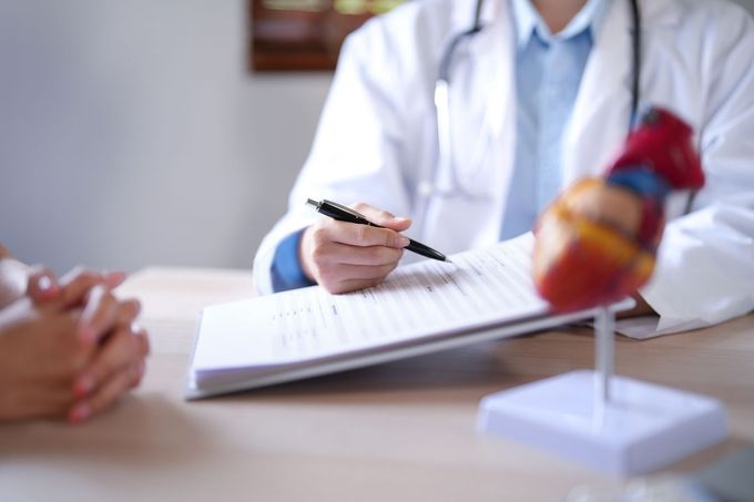 Un medico in camice bianco scrive su un blocco durante una visita, con un modello anatomico del cuore sulla scrivania.
