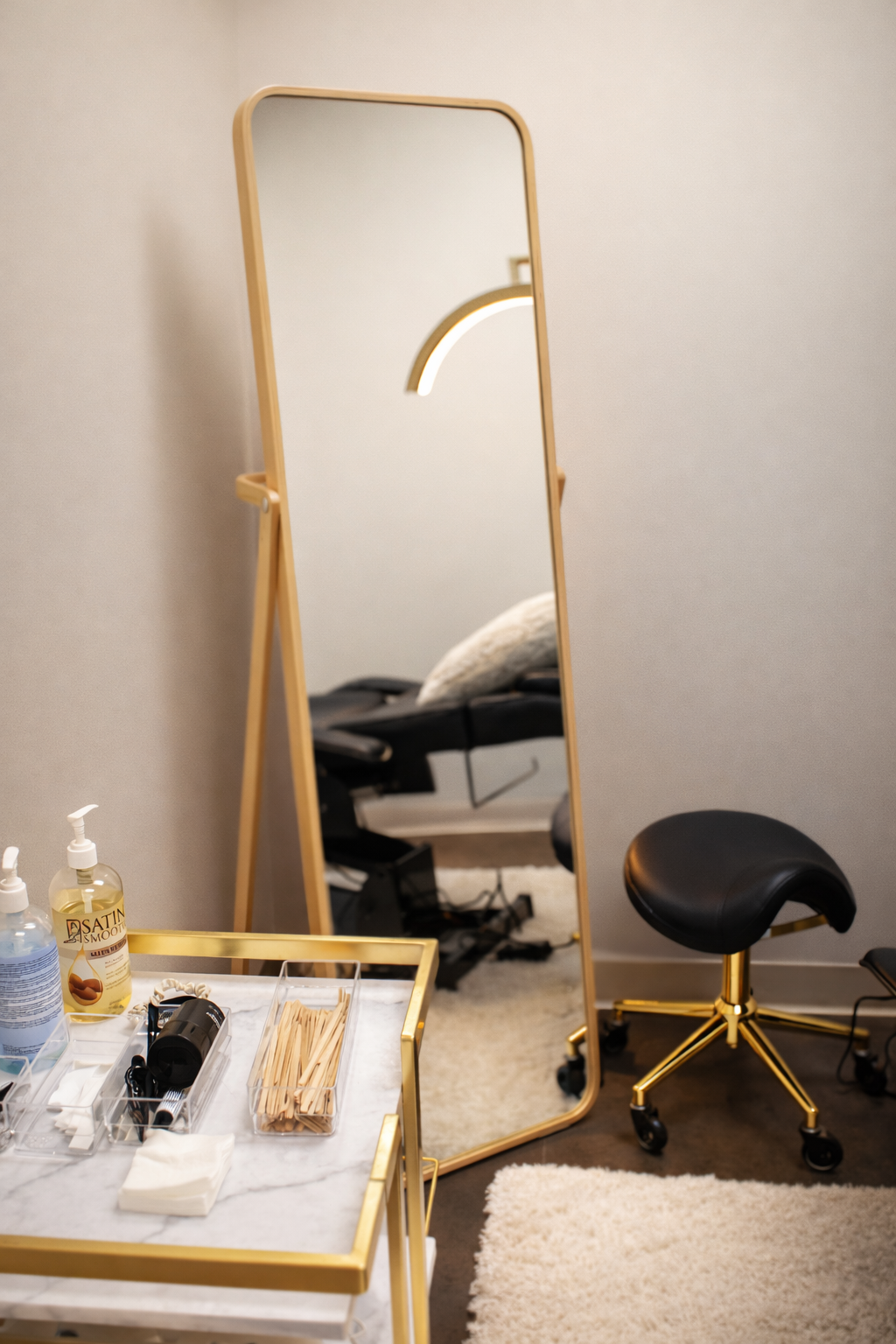 A gold-framed floor mirror stands beside a rolling cart and a black ergonomic stool in a bright, modern studio space.