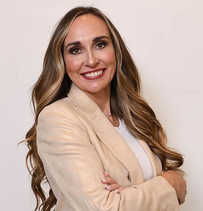 A smiling person with long brown hair wearing a light tan blazer over a white shirt, posed with arms crossed.