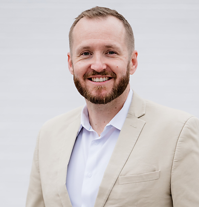 A person with a beard and light brown hair, wearing a beige blazer over a white collared shirt, smiling against a gray.