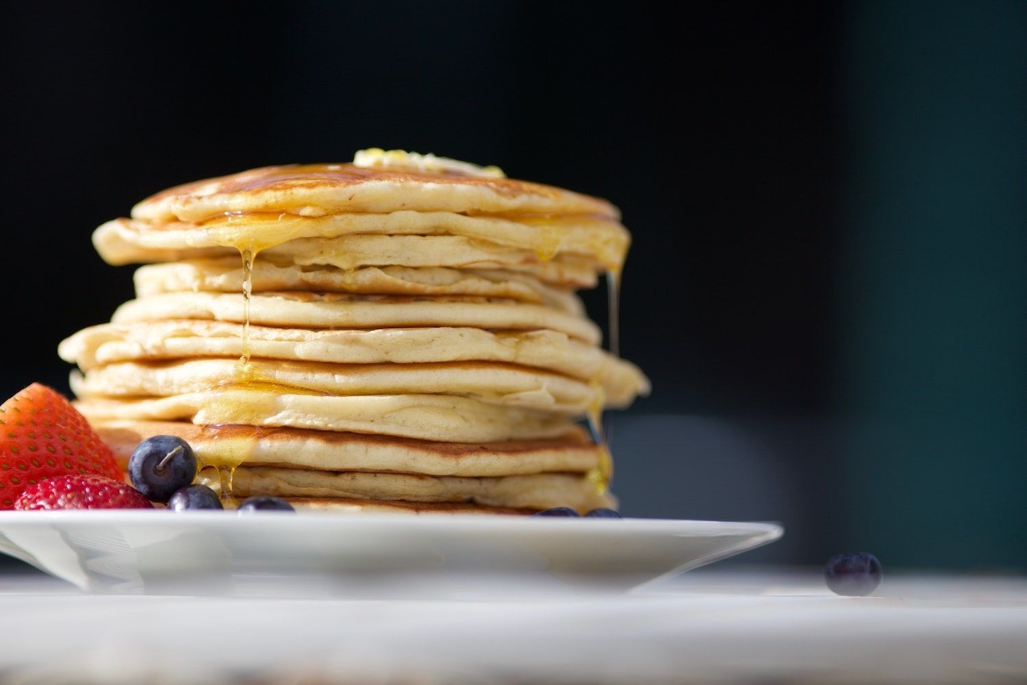 Stack of pancakes with syrup drizzling, served with strawberries and blueberries on a white plate.
