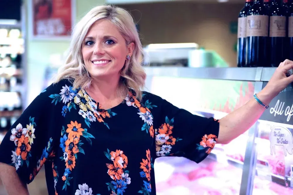 Woman in floral top smiles, leaning on a display case in a store.