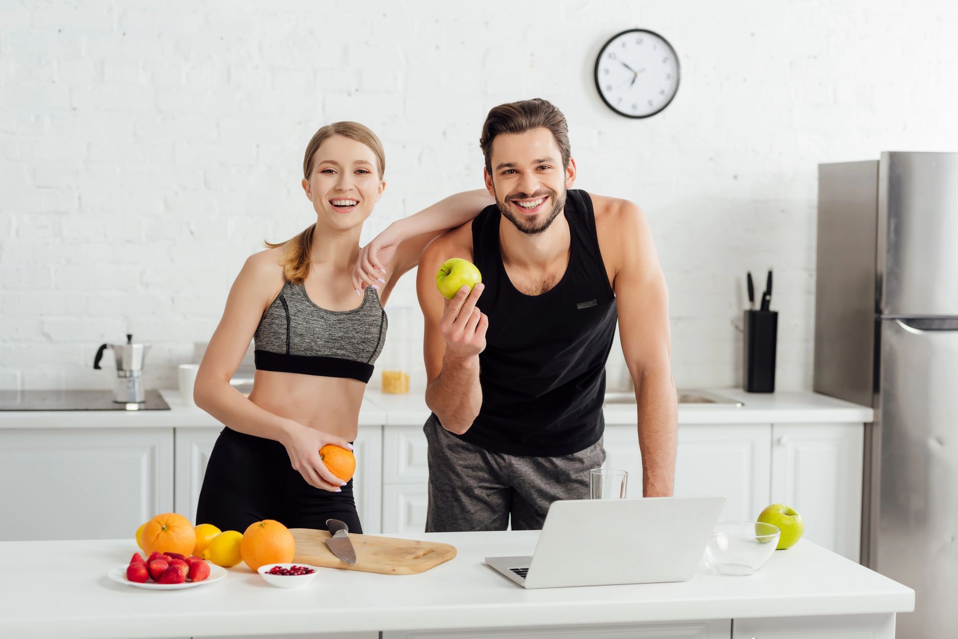 Couple in athletic wear in kitchen with fruits and laptop. Woman holds orange, man holds apple.