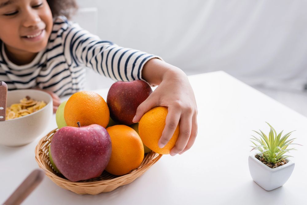 Child reaching for an orange from a fruit basket on a white table.