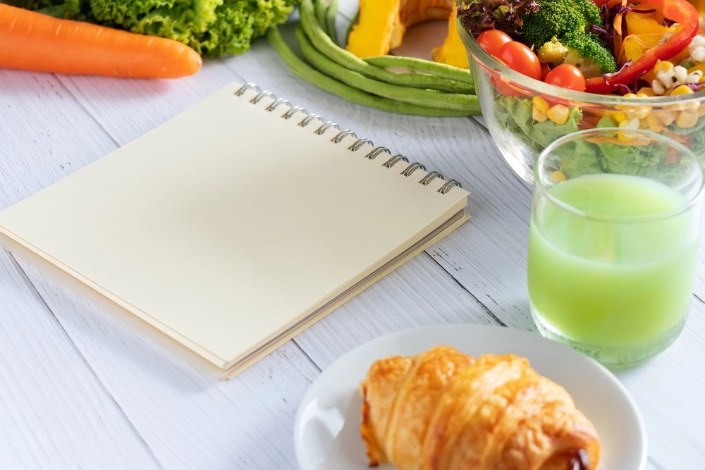 Notebook, vegetables, croissant, and juice on a white table. Healthy food and meal planning.