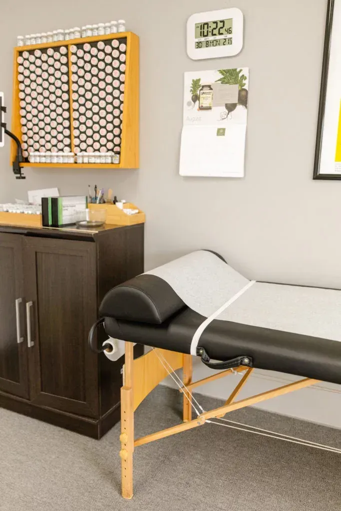 A massage table in a dimly lit office. Wooden cabinet with shelves of medicine bottles. A digital clock hangs on the wall.