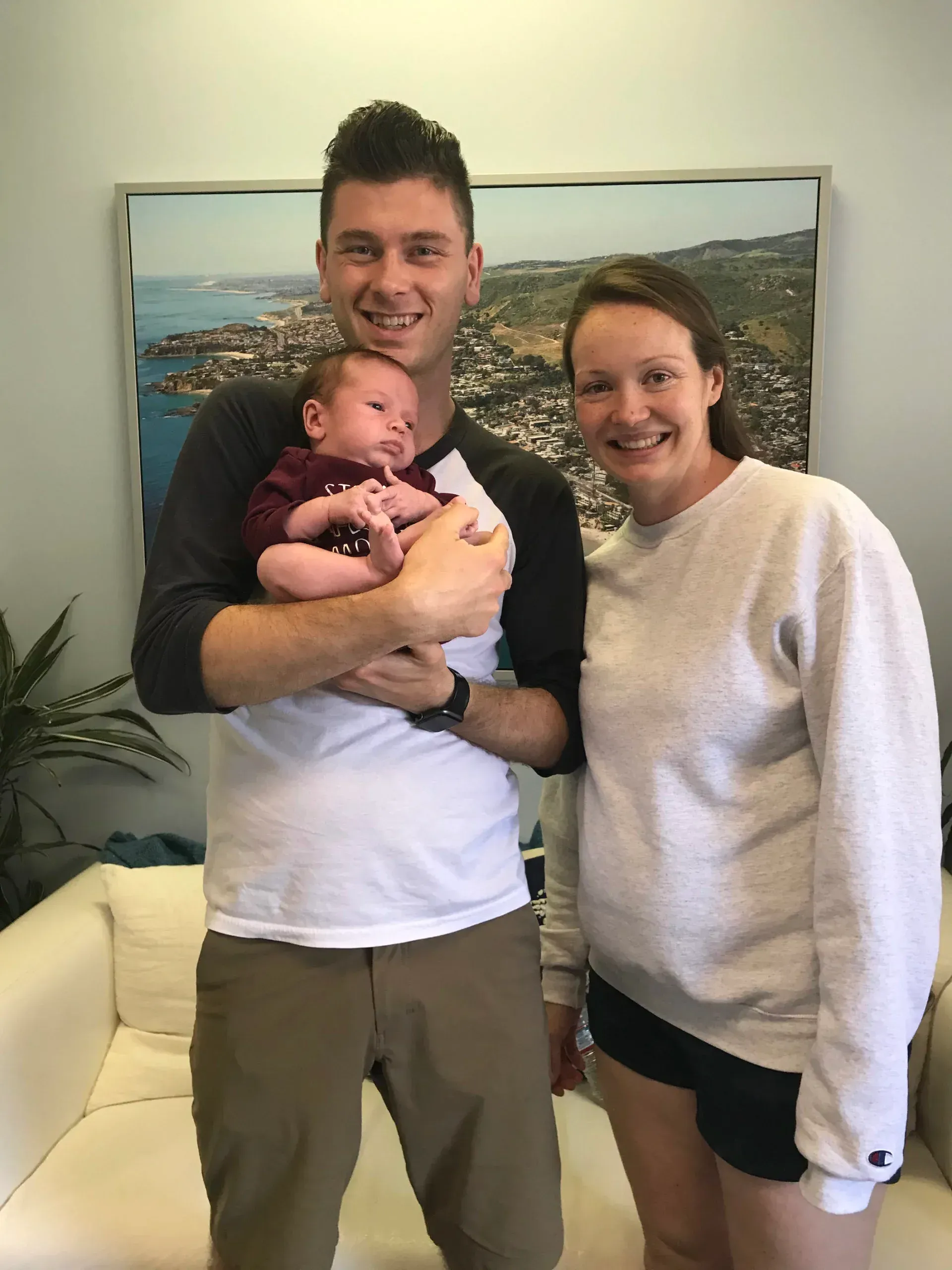 A man holds a baby; a woman stands beside them. They are smiling in a room with a painting on the wall.