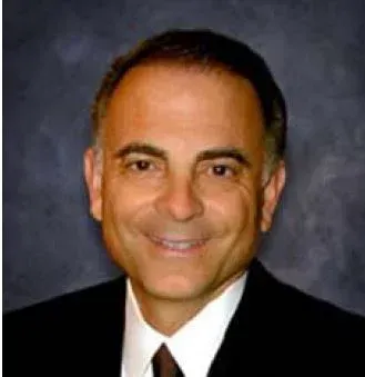 Smiling man in a black suit and white shirt, against a dark background.