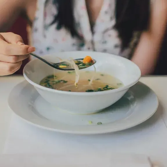 Woman eating soup from a white bowl, holding a spoonful with sprouts and vegetables.
