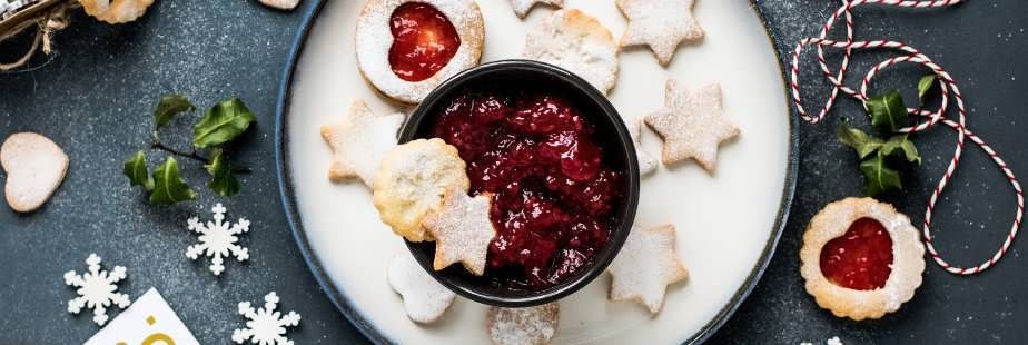 Cookies and jam on a plate. Star and heart-shaped cookies. Dark red jam in a black bowl. Dark, textured surface.