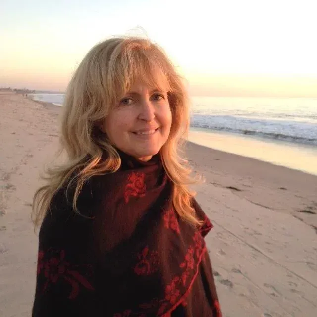 Woman with blonde hair, wrapped in a black shawl with red floral design, smiles on a beach at sunset.