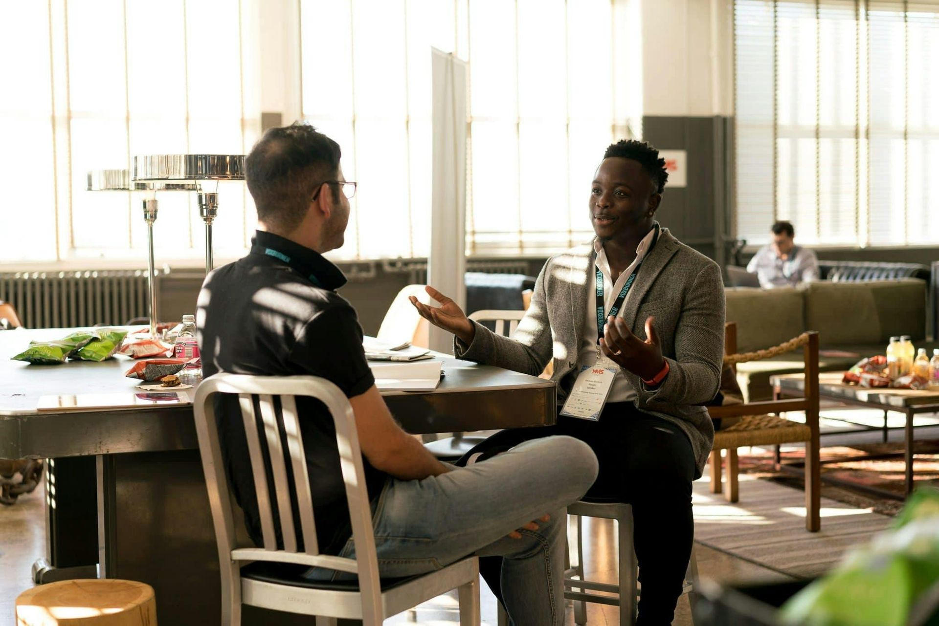 Two people talking at a café table by a bright window, with drinks and papers between them.