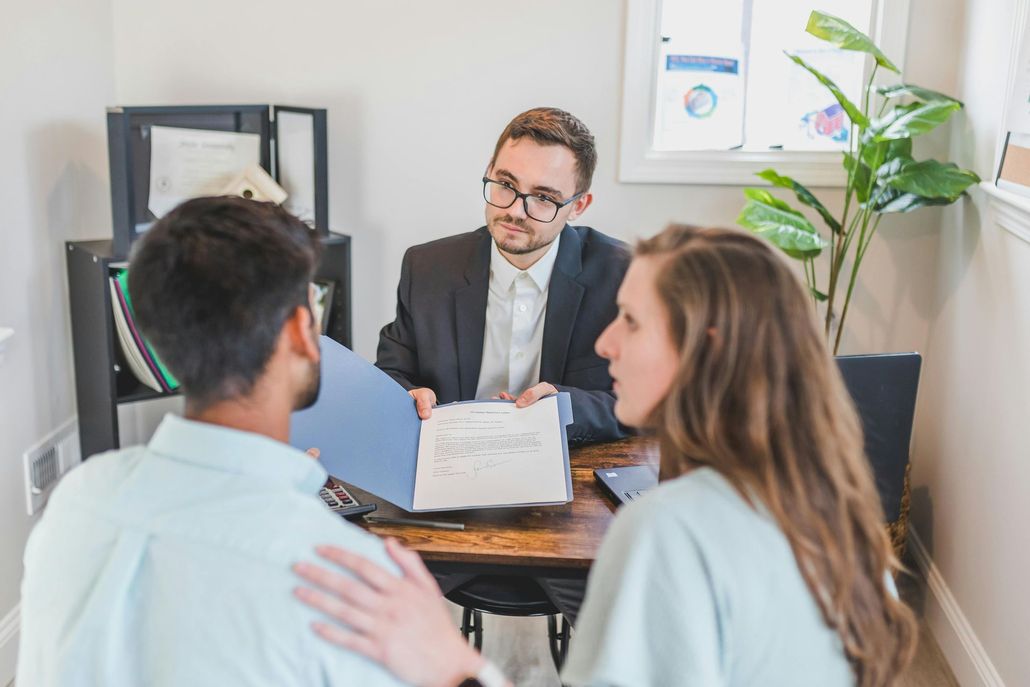 Three people in a small office meeting, discussing documents across a desk.
