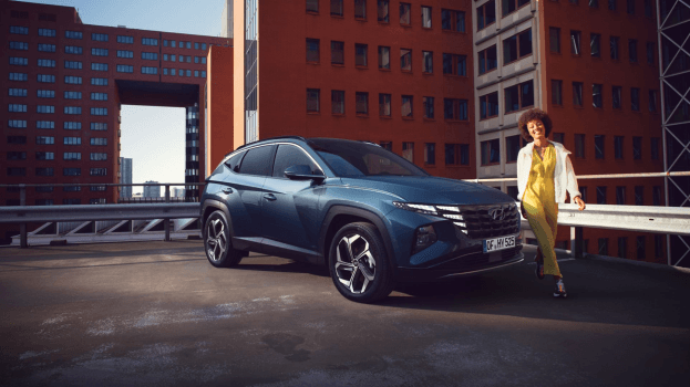 A woman is standing next to a blue car in a parking lot. Tucson