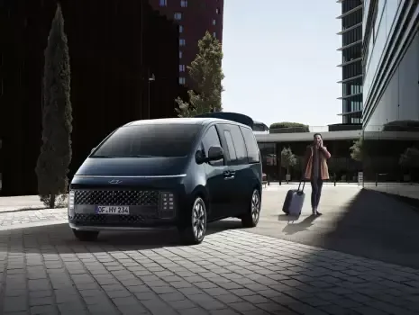 A woman is walking towards a blue minivan with a suitcase. Staria