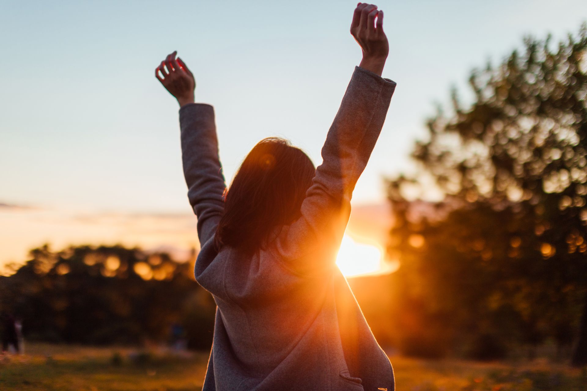 A woman is standing in a field with her arms in the air at sunset.