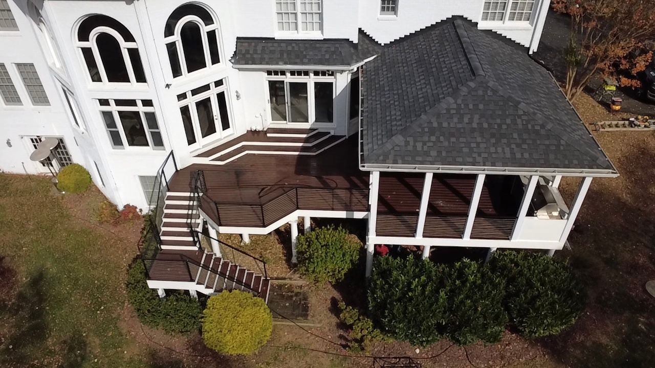 White house with tiered wooden deck and screened-in porch, surrounded by green shrubs and grass.