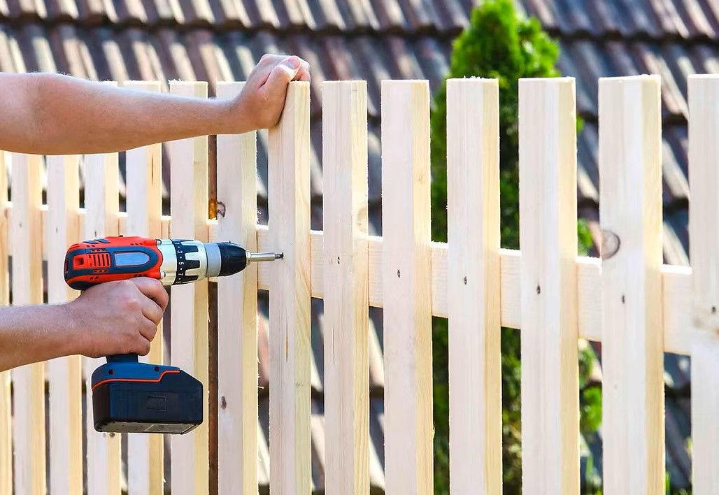 Person using a power drill to attach wooden planks to a fence in an outdoor setting.