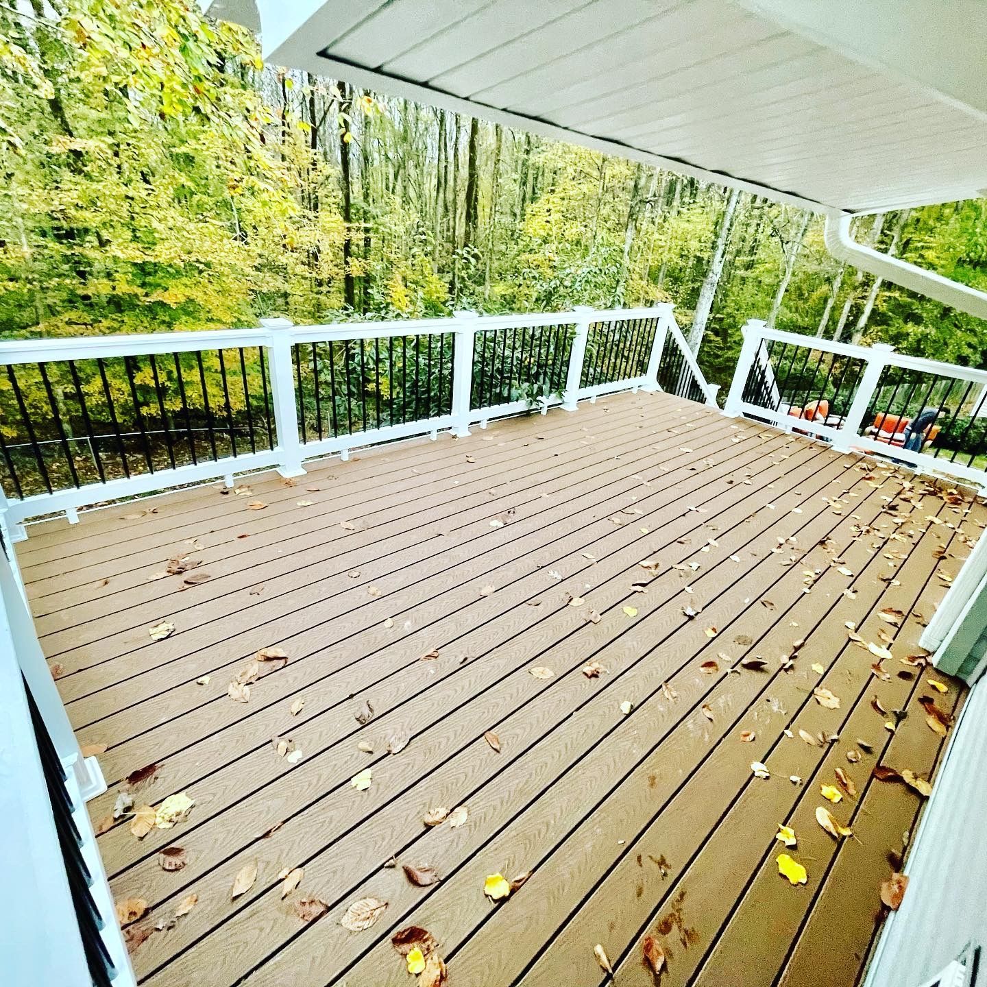 Brown wooden deck with fallen leaves, white railings, black balusters, and forest in background.