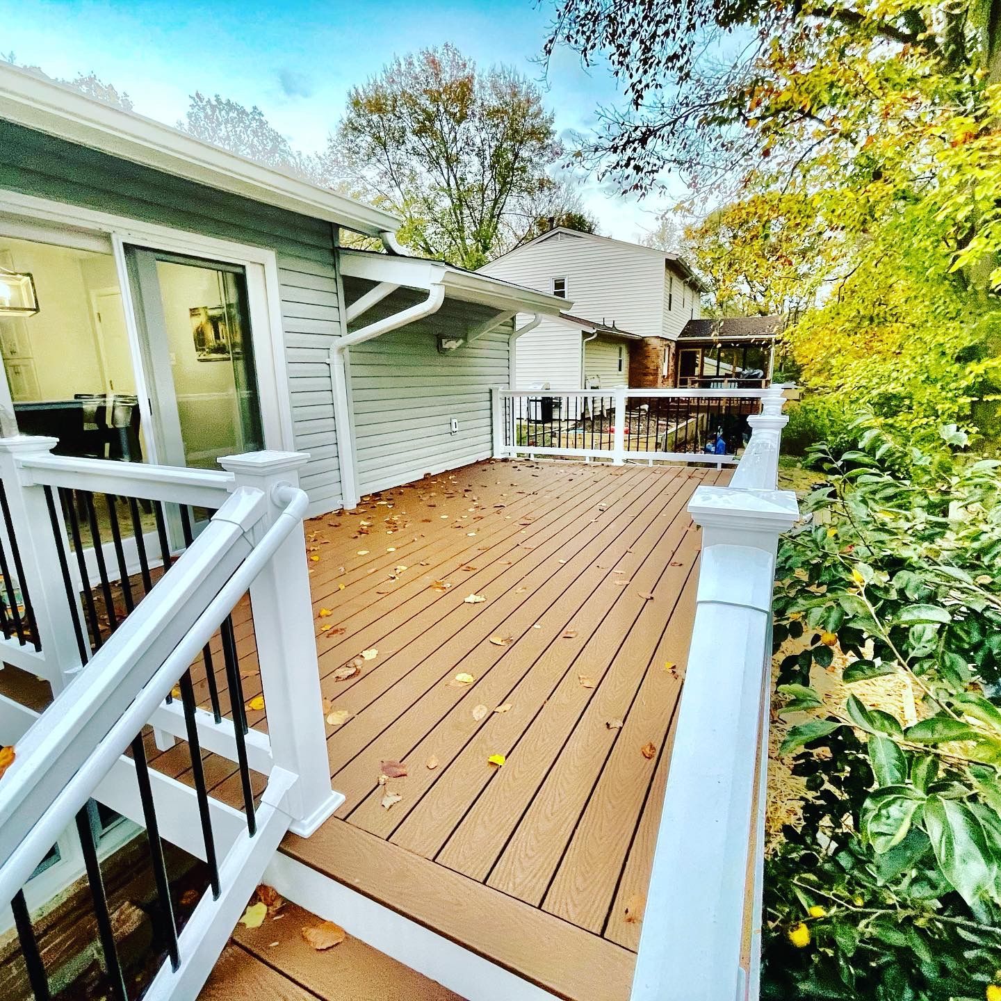 Wooden deck with white railing, black spindles, and fallen leaves. View of a house with trees in the background.