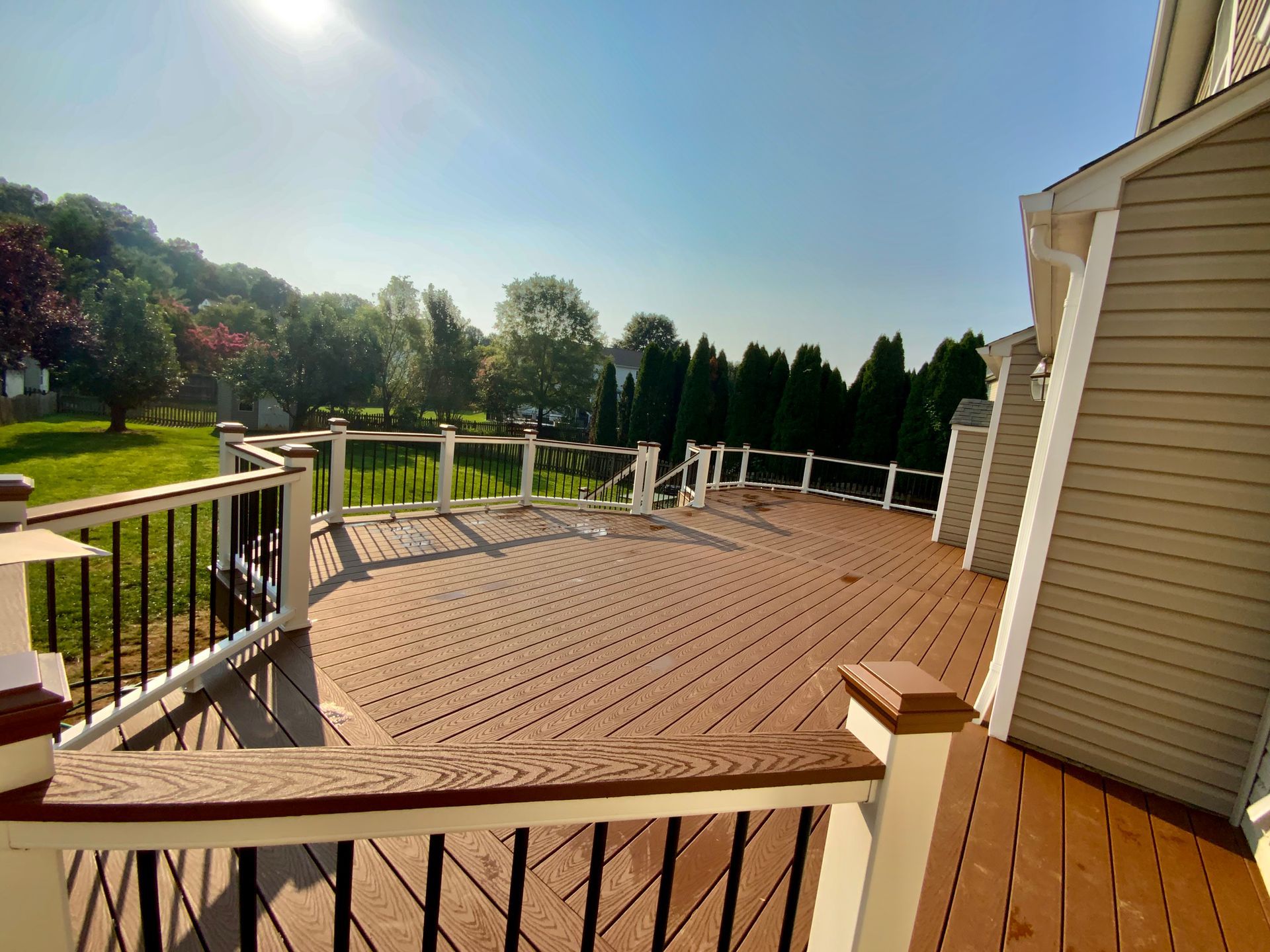Wooden deck with brown and white railings, overlooking a green lawn and trees under a sunny blue sky.