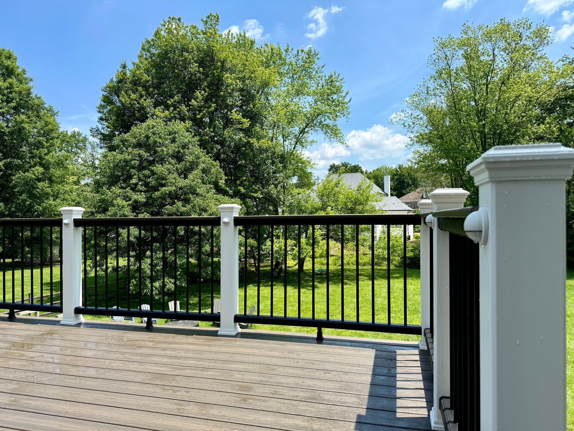 View from a deck with black railing and white posts, overlooking a green yard and trees under a blue sky.